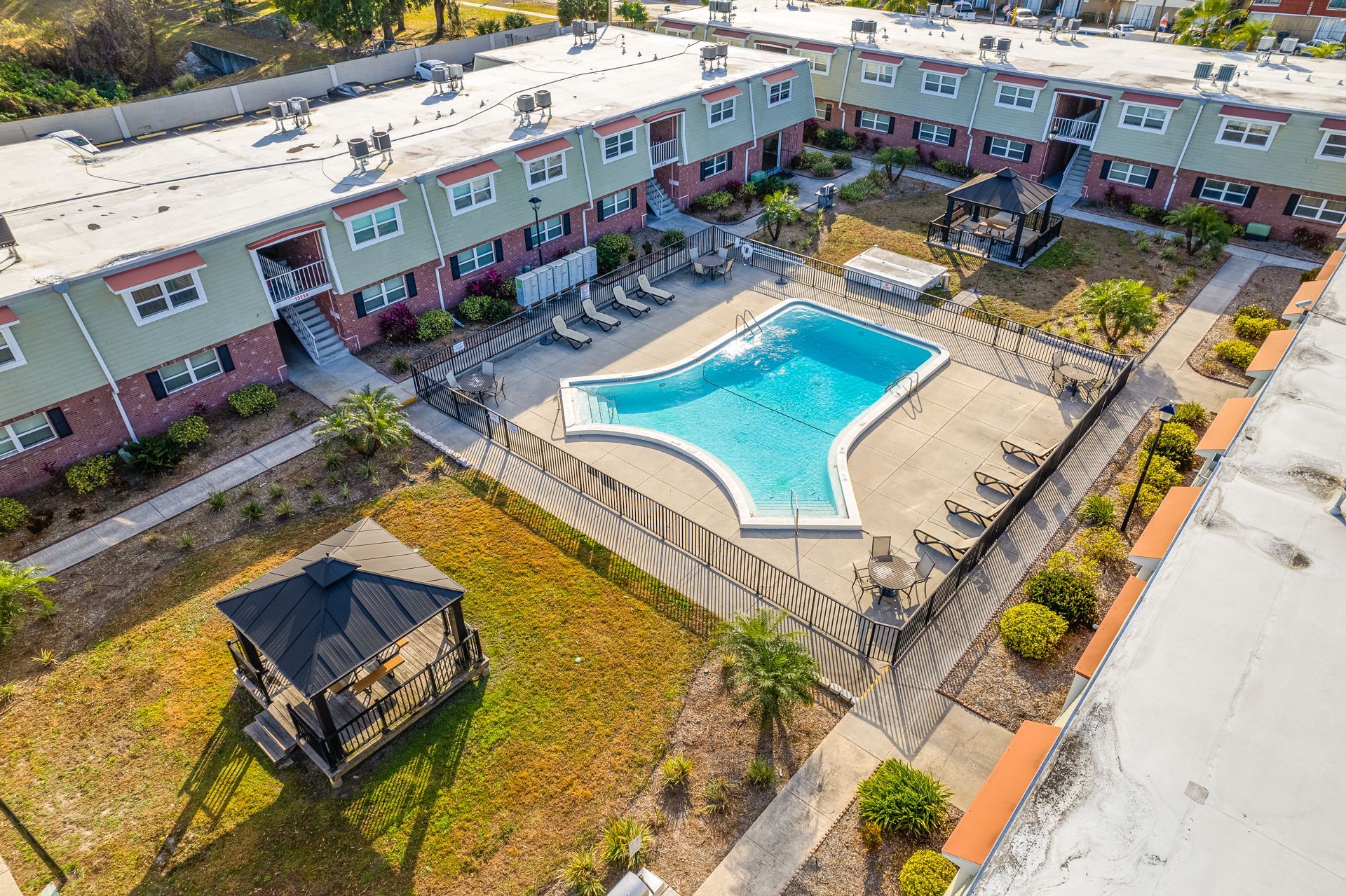 Aerial view of apartment complex with a pool, gazebo, and sun loungers. Buildings have red brick and green trim.