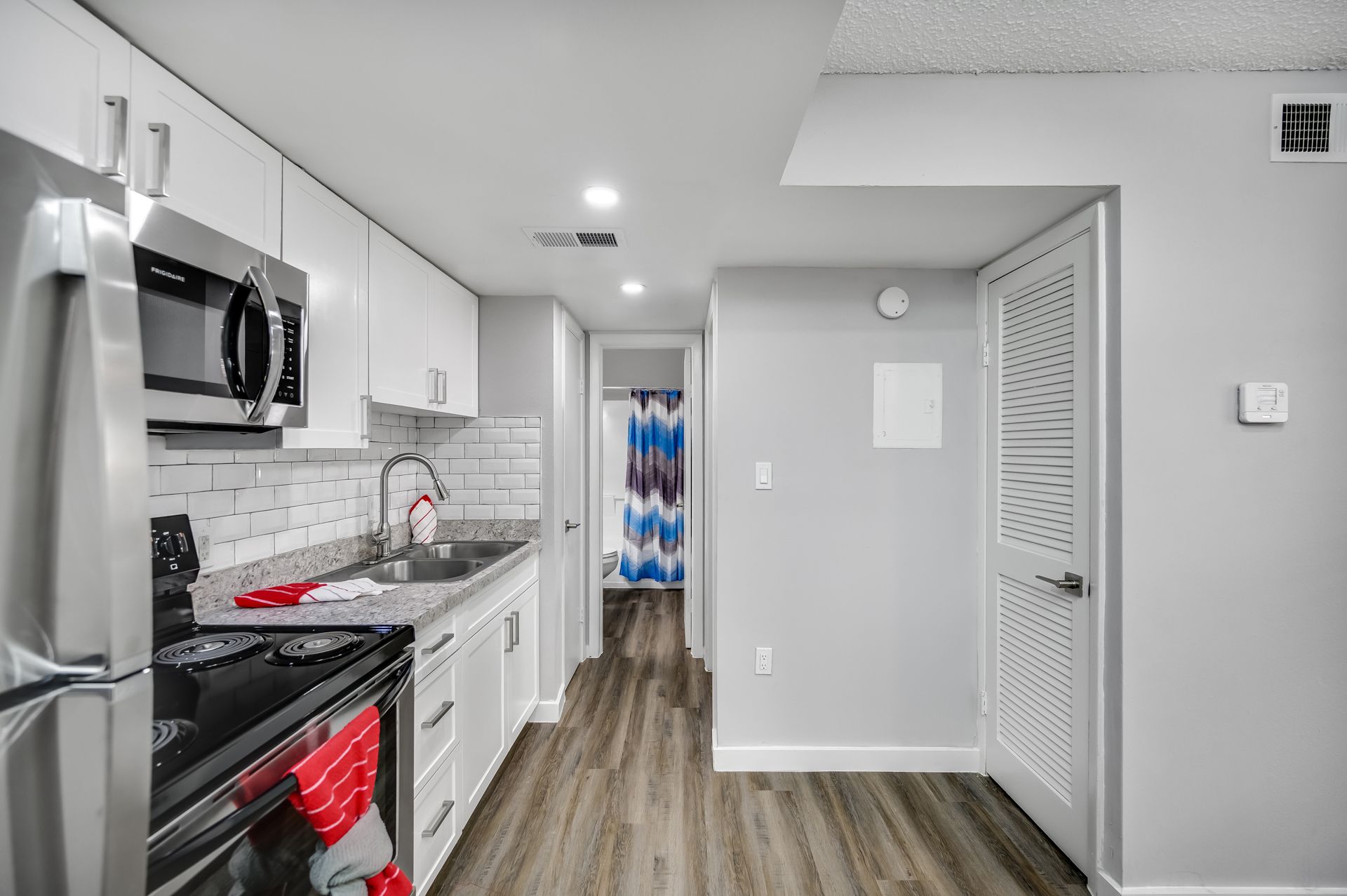 Modern kitchen with white cabinets, stainless steel appliances, and gray countertops, leading to a hallway with a bathroom.