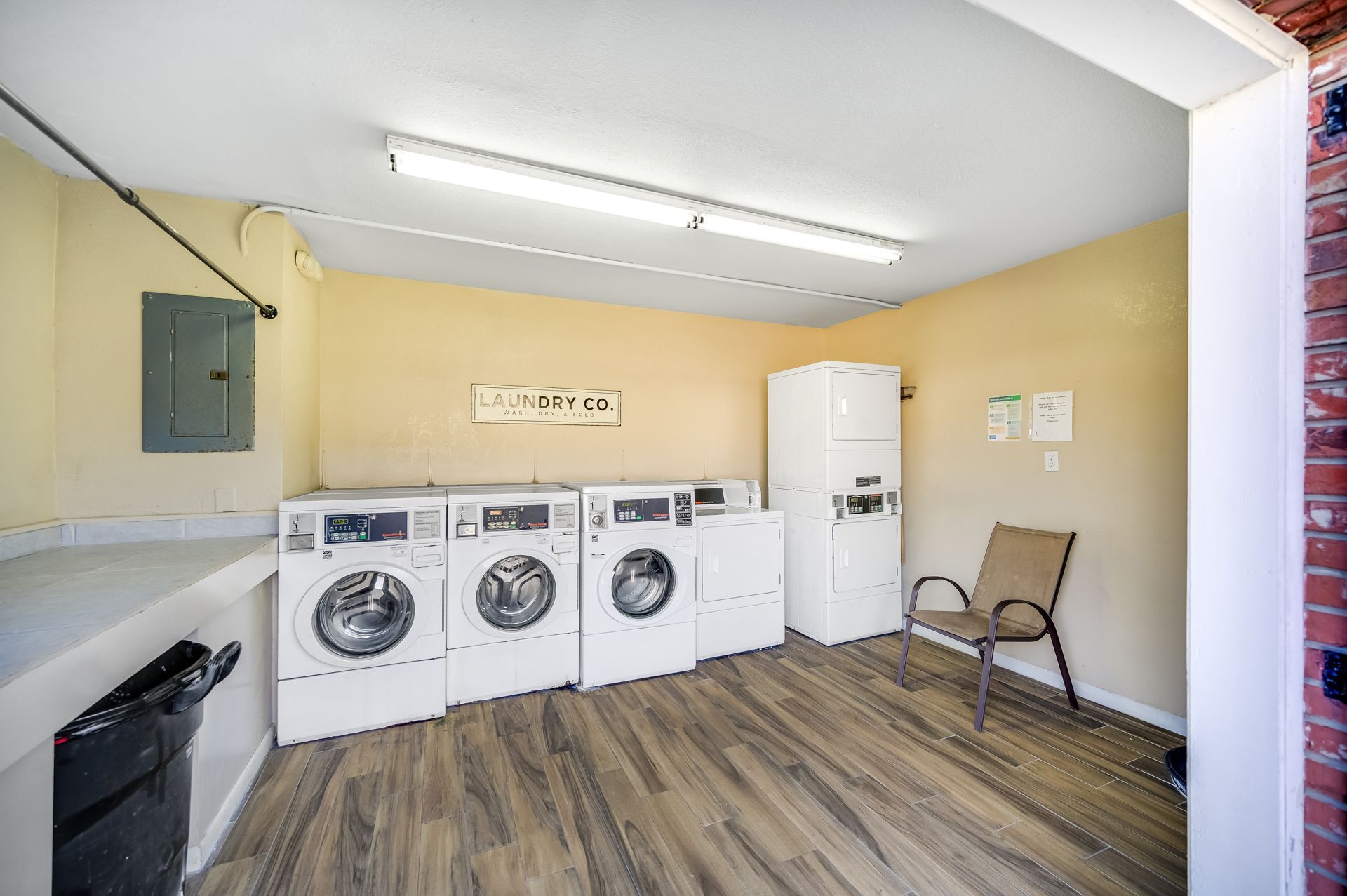 Laundry room with washing machines, a dryer, and a chair. Beige walls, wood-look floor, and a black trash bag.