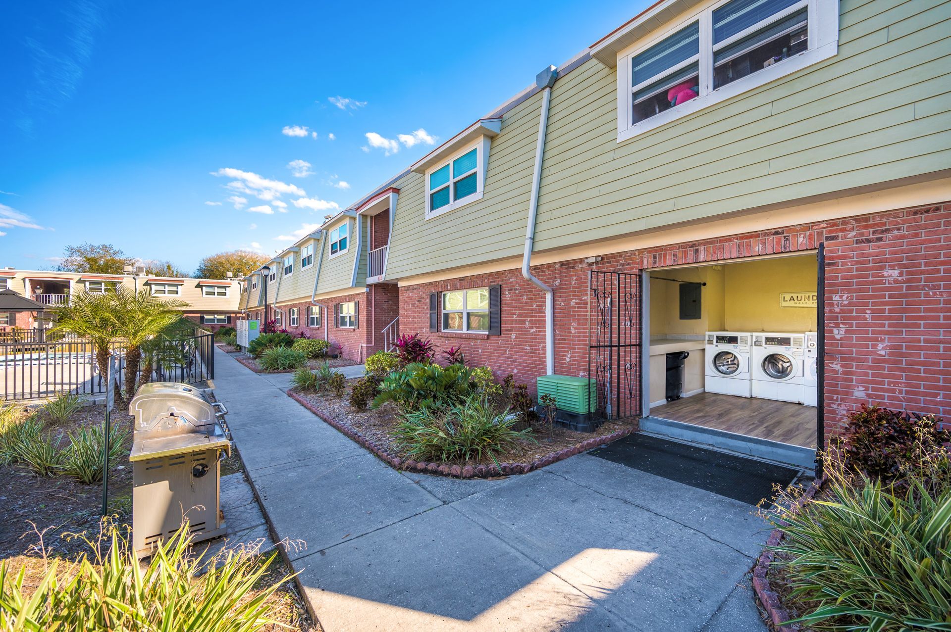 Apartment complex exterior with brick and siding, a laundry area, and a walkway under a blue sky.