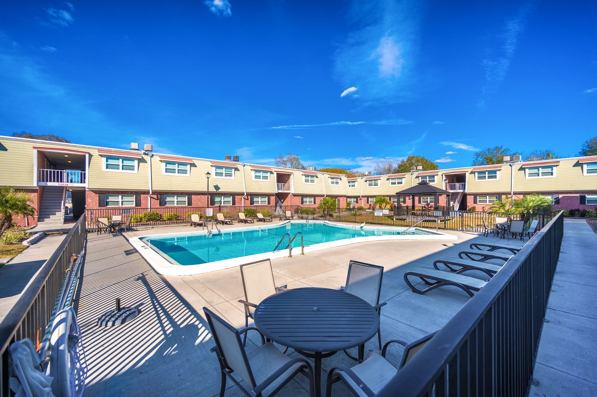 Swimming pool and lounge area surrounded by a two-story apartment complex under a blue sky.