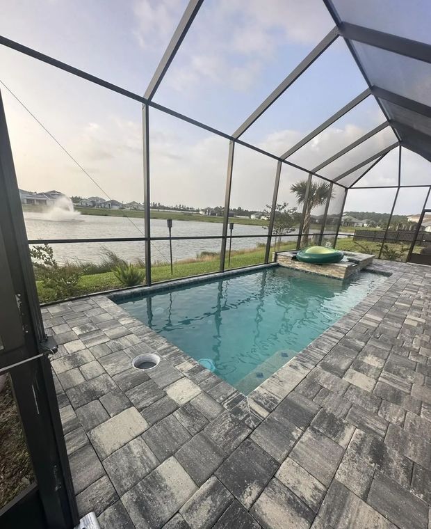 A swimming pool area near Flagler Beach, FL, with its surfaces restored to look brand new again by Top Elite Properties featuring perfectly sealed and restored grey stone pavers, inside a screened-in lanai overlooking a lake with a fountain.