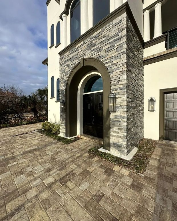 An arched stone entryway with a dark door, flanked by outdoor lanterns and set against a pristine pressure washed paved driveway at sunset in Palm Coast, Flagler County, FL.
