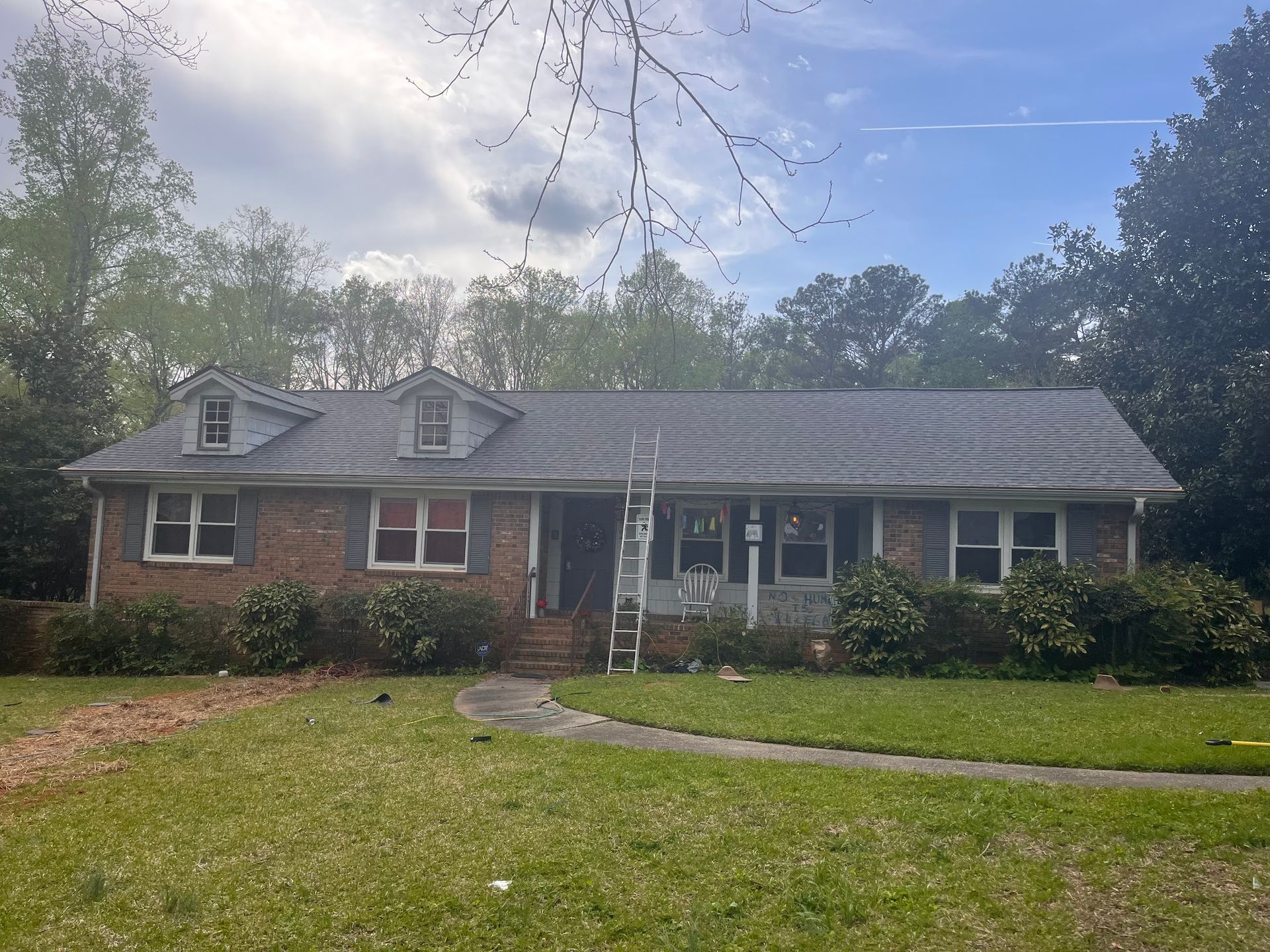 Brick house with dark roof and ladder, surrounded by green grass, bushes, and trees under a cloudy sky.