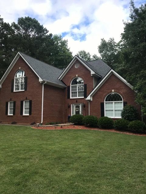 Brick house with black shutters, arched windows, and a dark roof, set on a green lawn with trees in the background.