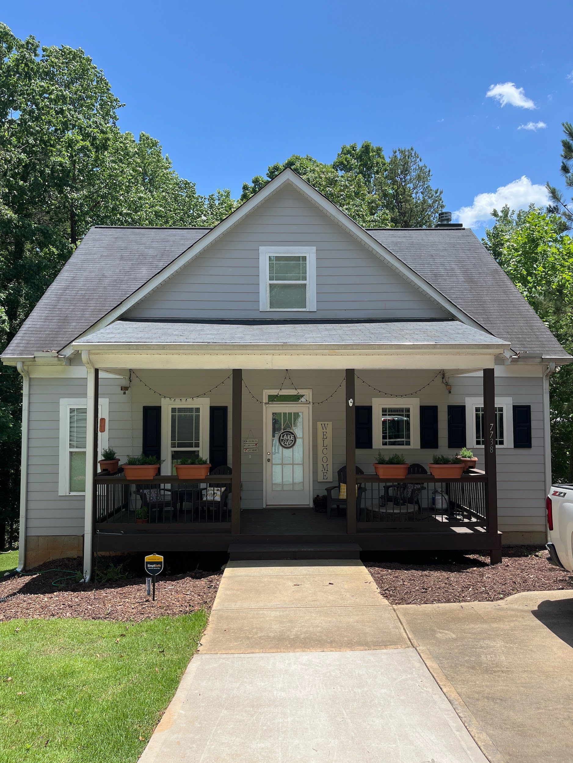 A light grey, gabled house with a porch, black shutters, and a concrete walkway under a blue sky.