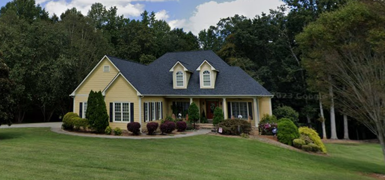 Yellow house with black roof and dormers, on a green lawn with trees in the background.