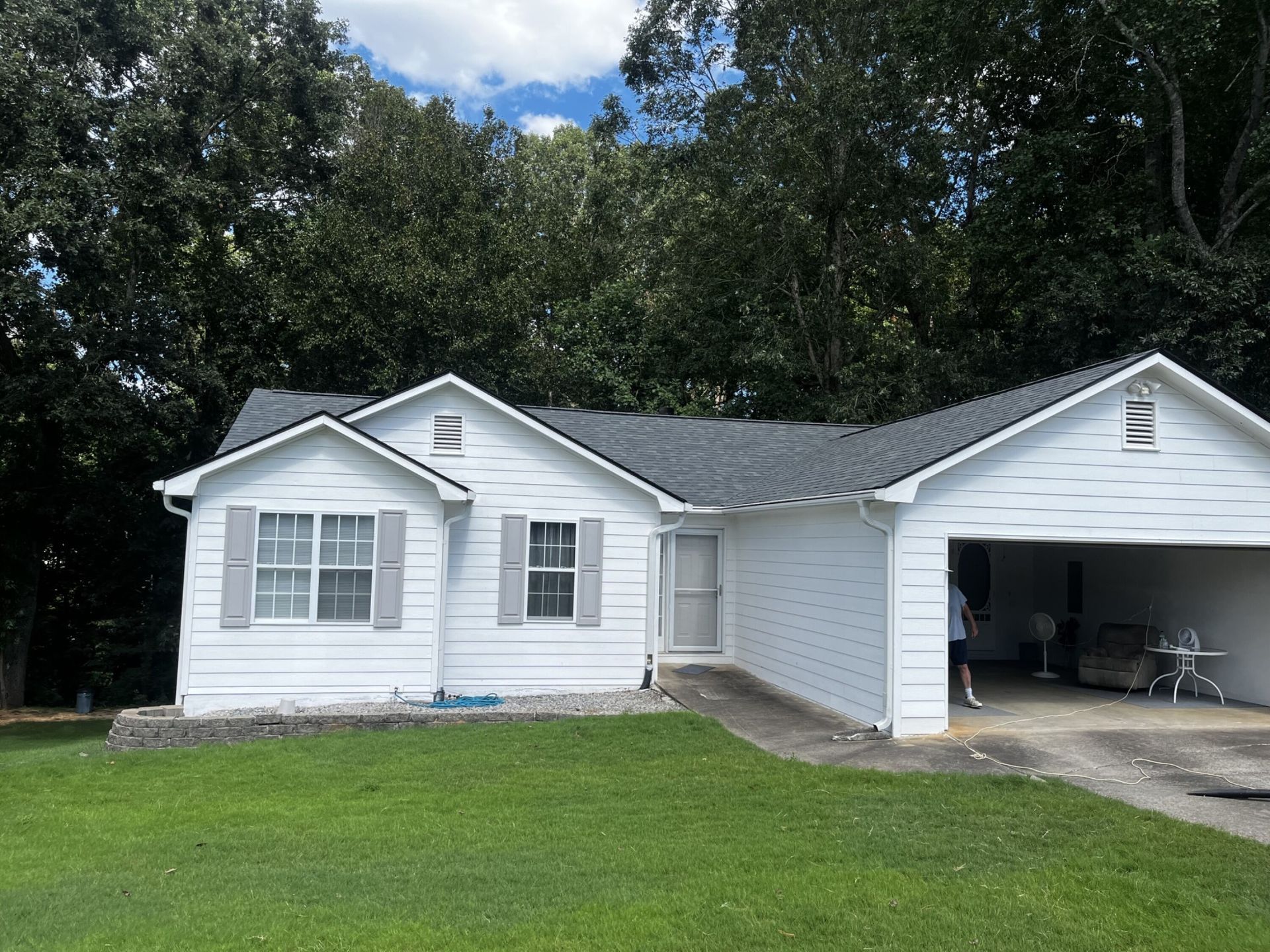 White house with gray roof, small lawn, and carport. Trees in the background, blue sky.