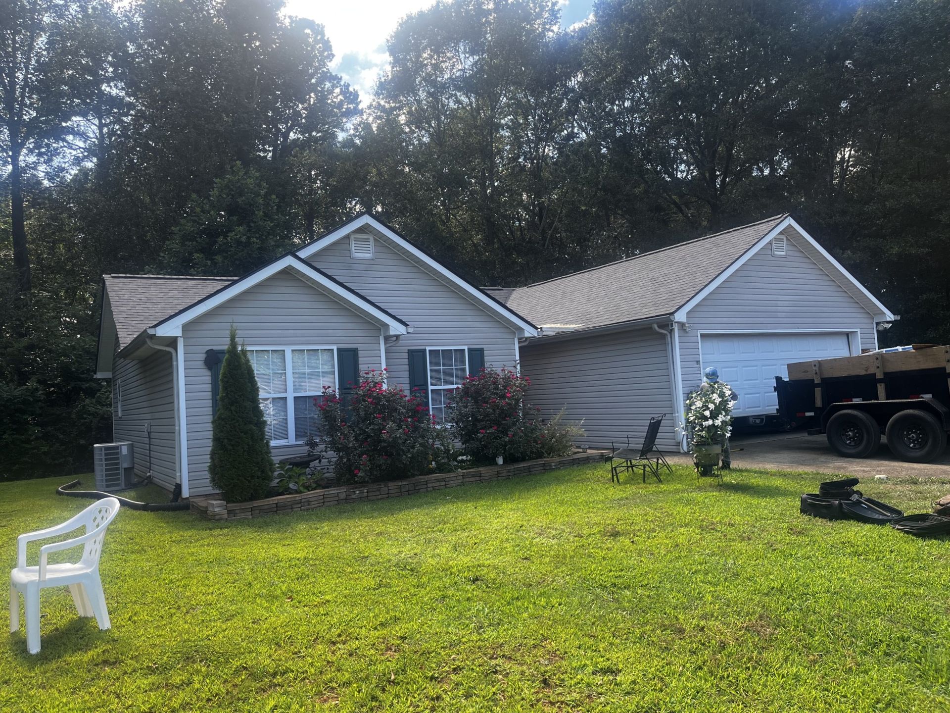 Gray house with a green lawn, bushes, and a tree in front. A white chair is on the left.