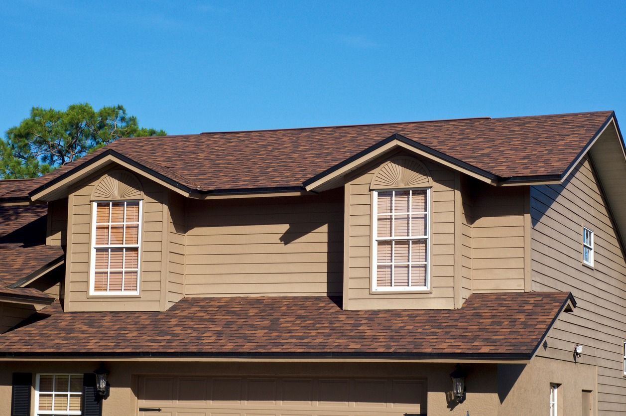 Tan house with brown roof and two dormer windows against a blue sky.