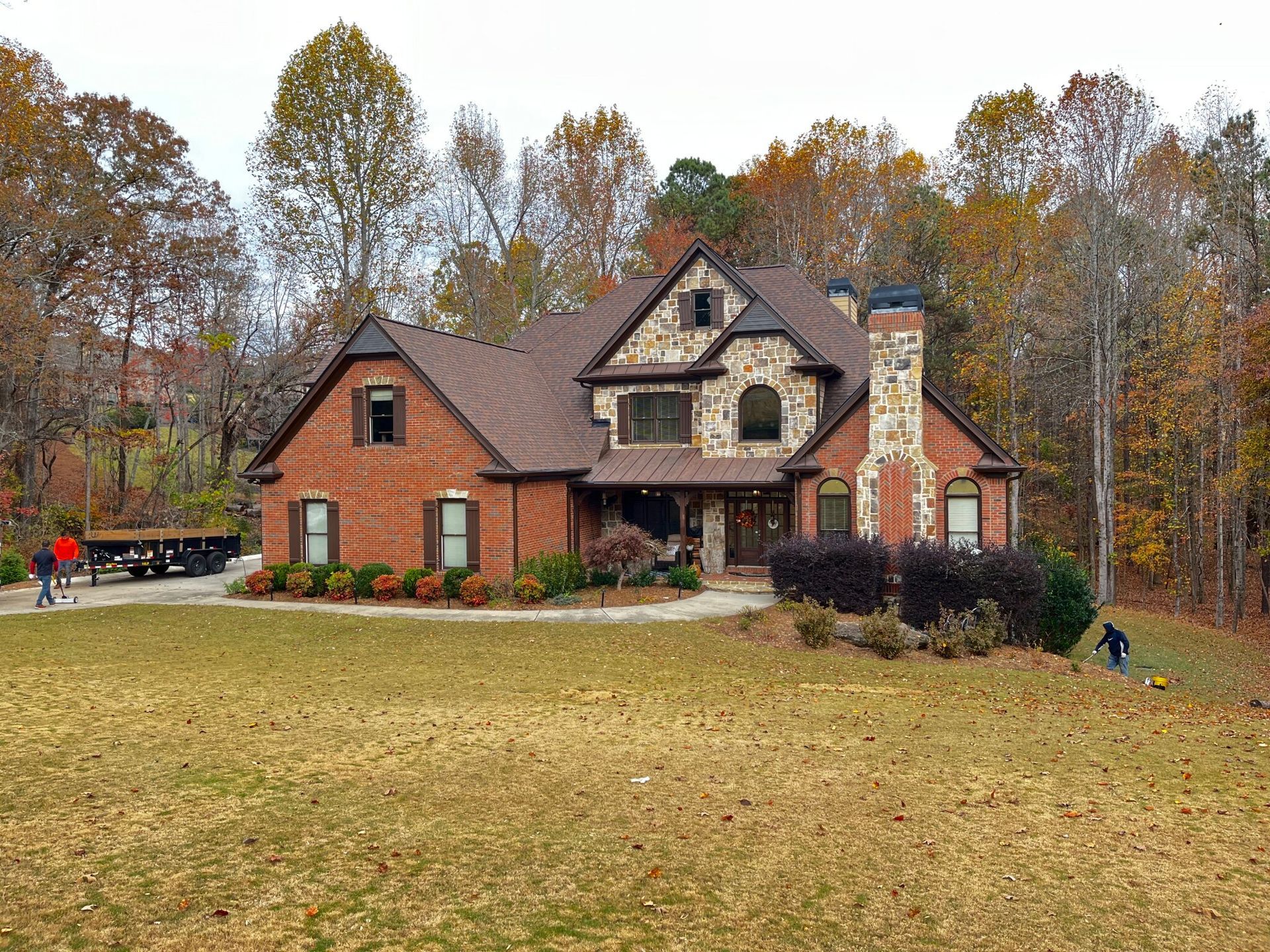 Brick house with brown roof, stone accents, and fall foliage in the background.