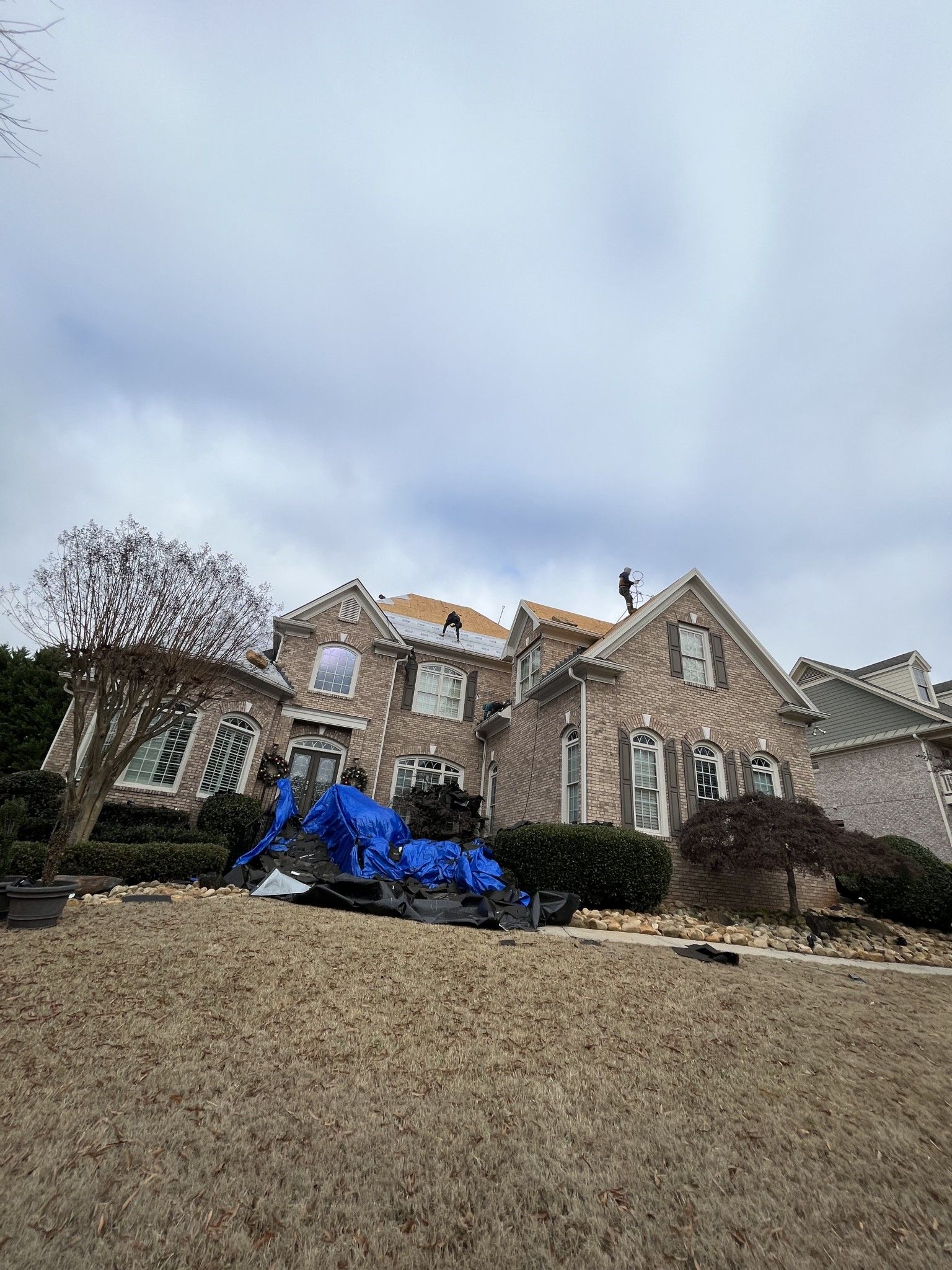 House under construction; roof being replaced, covered with blue tarps. Workers on the roof, cloudy sky.
