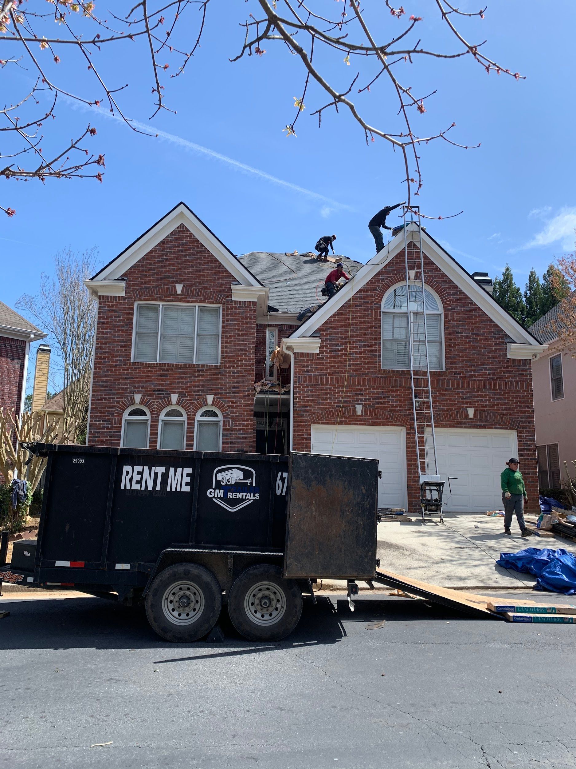 Roofers on a brick house replacing shingles. A dumpster trailer is parked in the driveway.