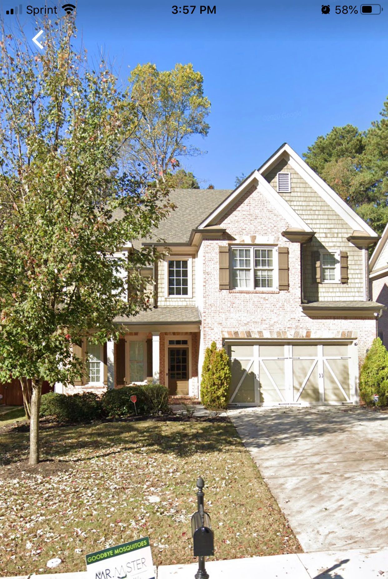 Two-story beige brick house with brown shutters and a two-car garage, located on a tree-lined lot.