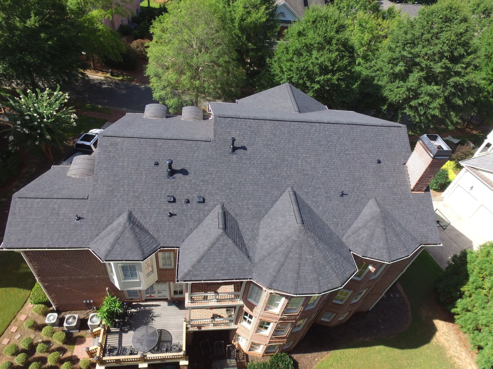 Overhead view of a house with a dark gray shingled roof, red brick siding, and surrounded by trees and lawn.