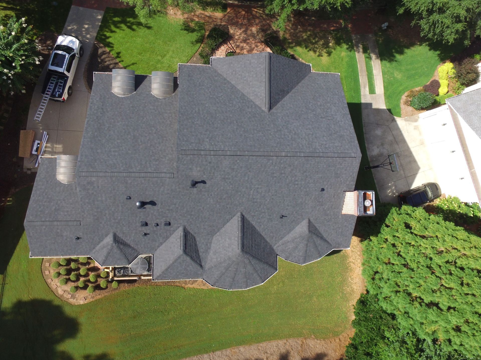 Overhead view of a house with a dark gray roof surrounded by green grass and trees. A car is parked in the driveway.