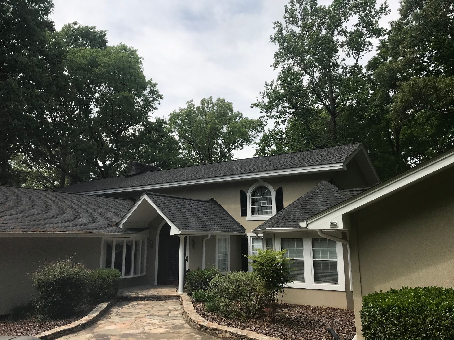 Beige house with black shingle roof, surrounded by trees.