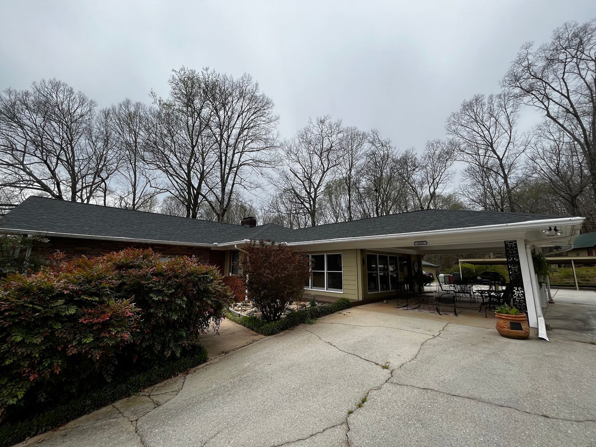 A ranch-style house with a carport and driveway under a cloudy sky. Trees line the background.