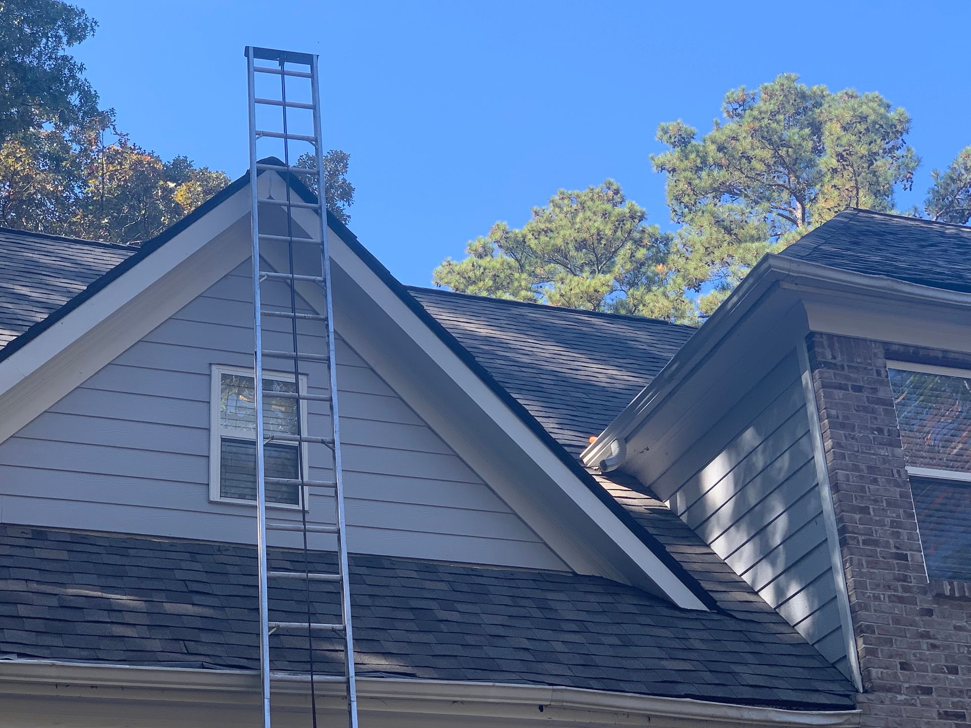 A ladder leaning against a house roof; blue sky and trees in the background.