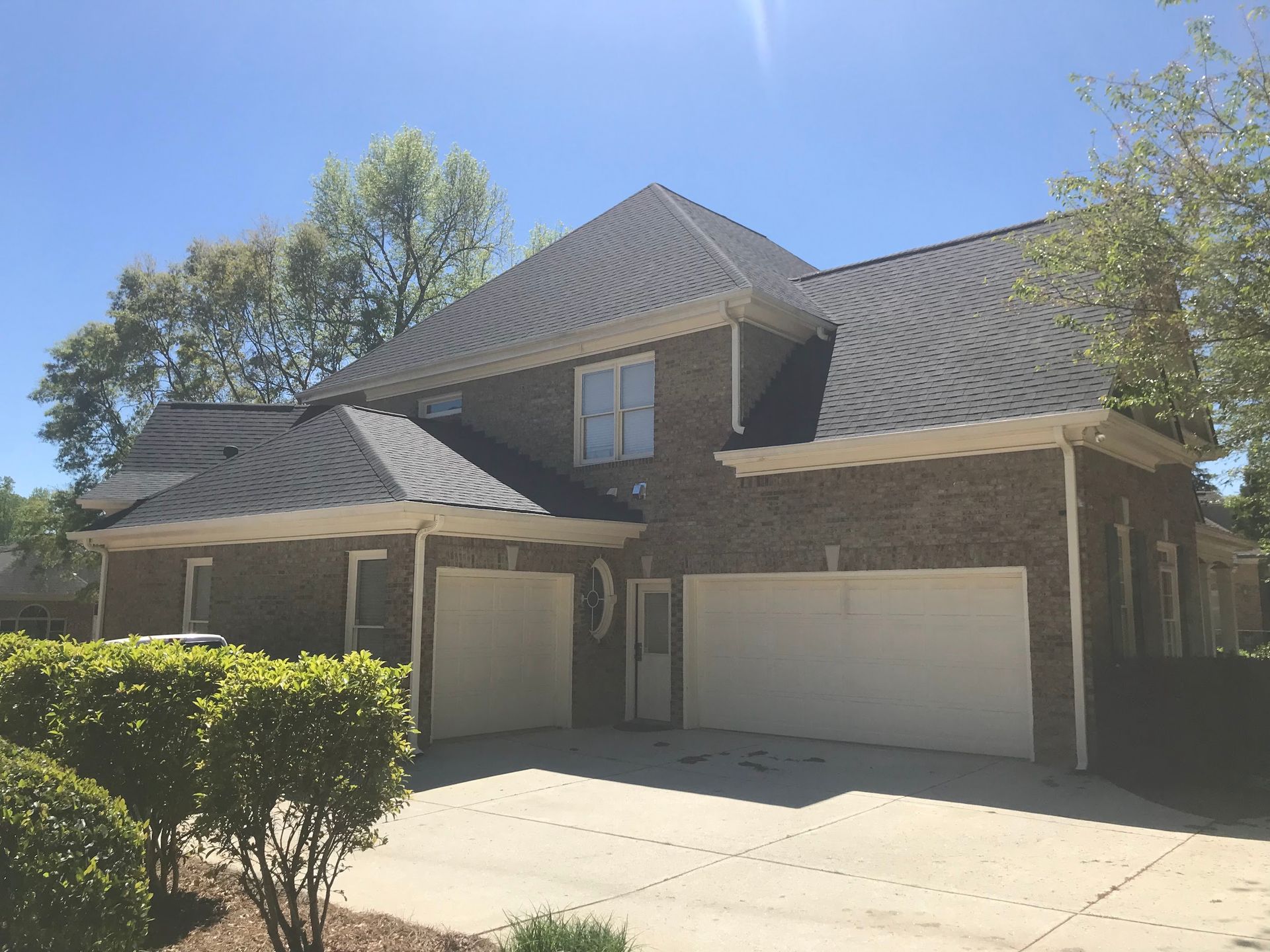 Two-story brick house with white garage doors and a dark shingled roof under a bright blue sky.