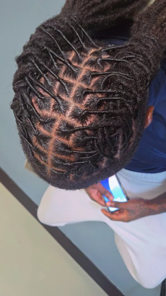 A top-down view of someone with neat, square-parted locs, wearing a dark shirt and light pants while holding a phone.