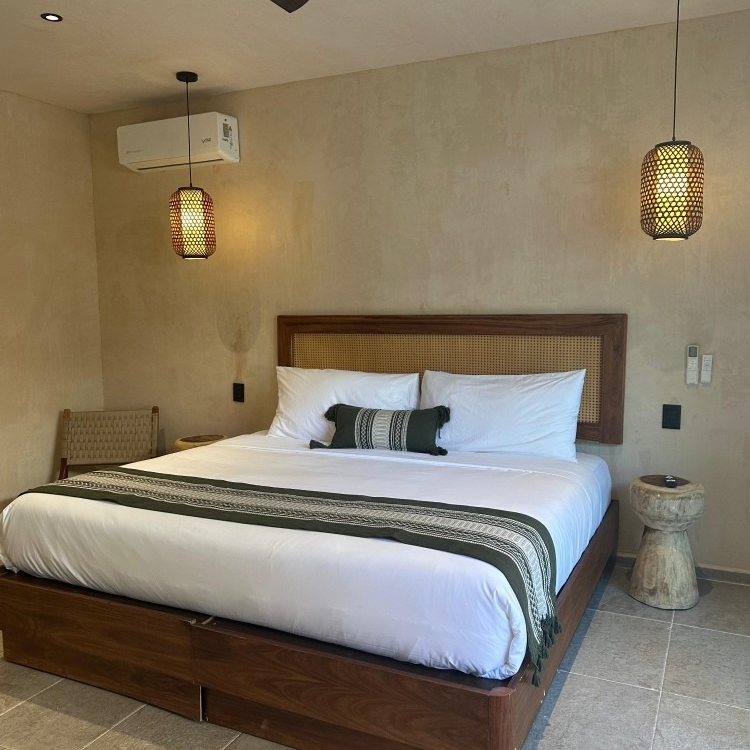 Bedroom with a bed, two pendant lights, and a side table. Neutral walls, wooden accents, and a green bed runner.