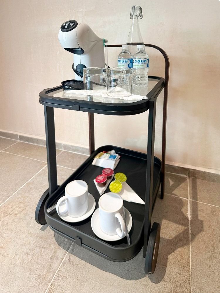 Black tea cart with coffee machine, water bottles, cups, and pods, on tile floor.