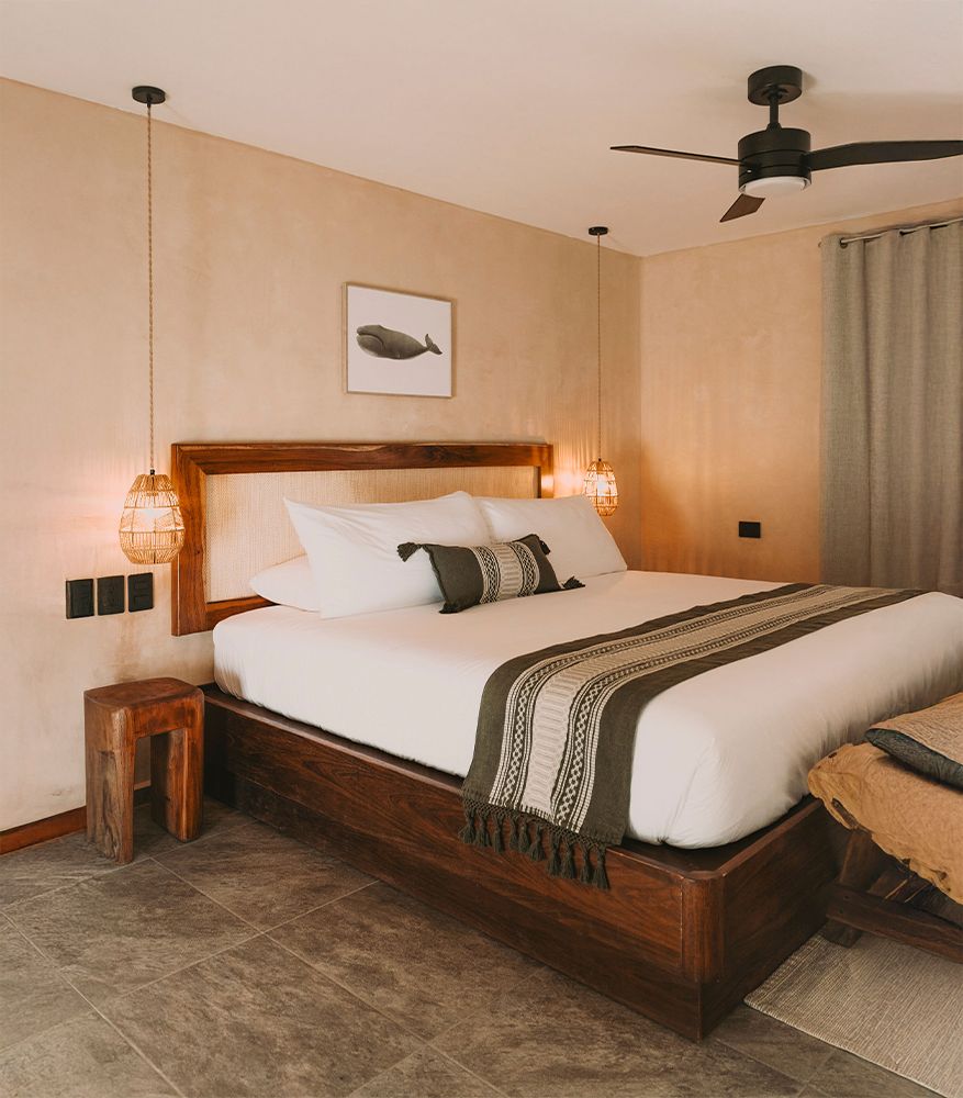 Bedroom with a wooden bed, woven headboard, white bedding, green runner, two hanging lamps, and neutral walls.