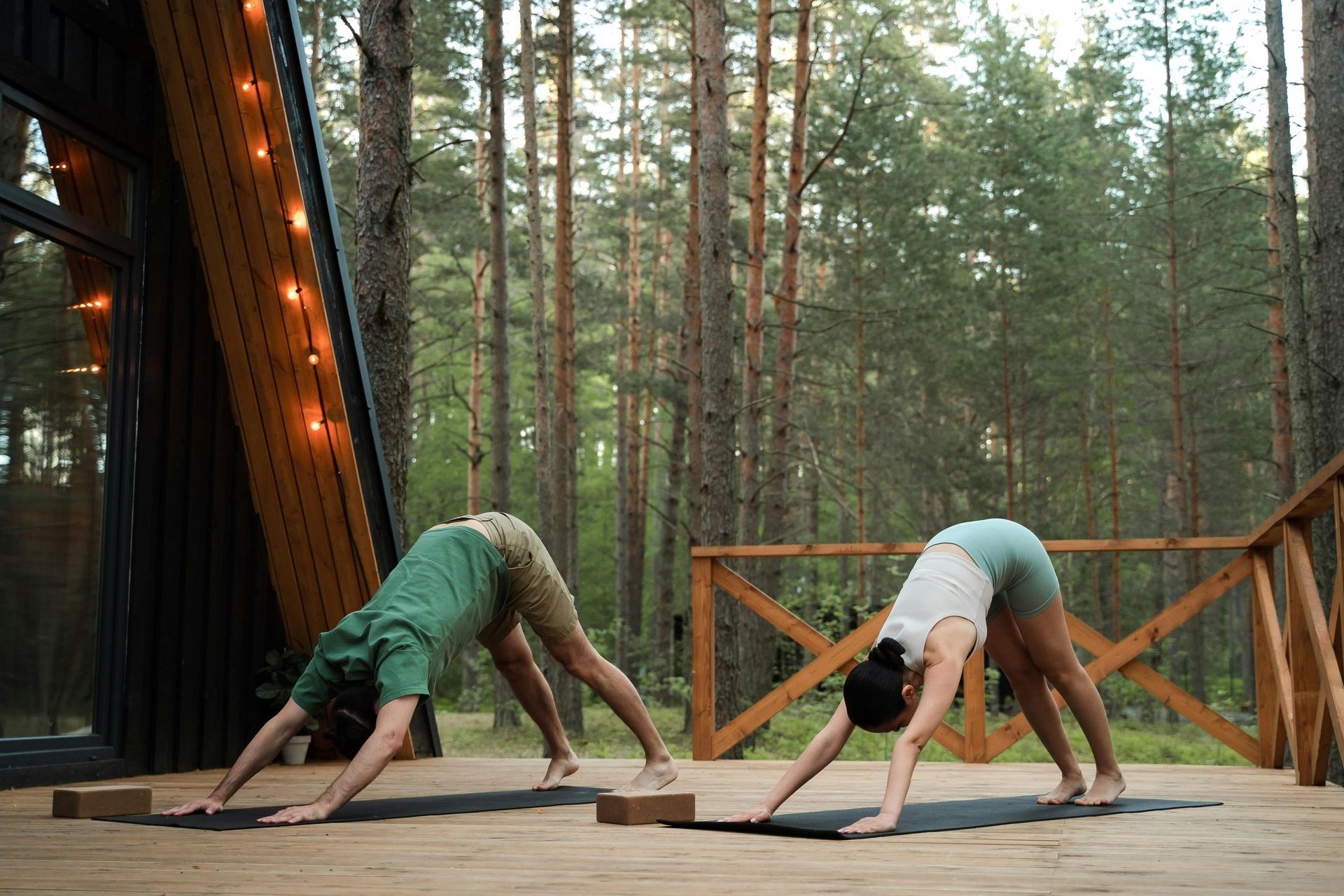 Two people doing yoga outdoors, downward-facing dog pose on a wooden deck in front of trees.