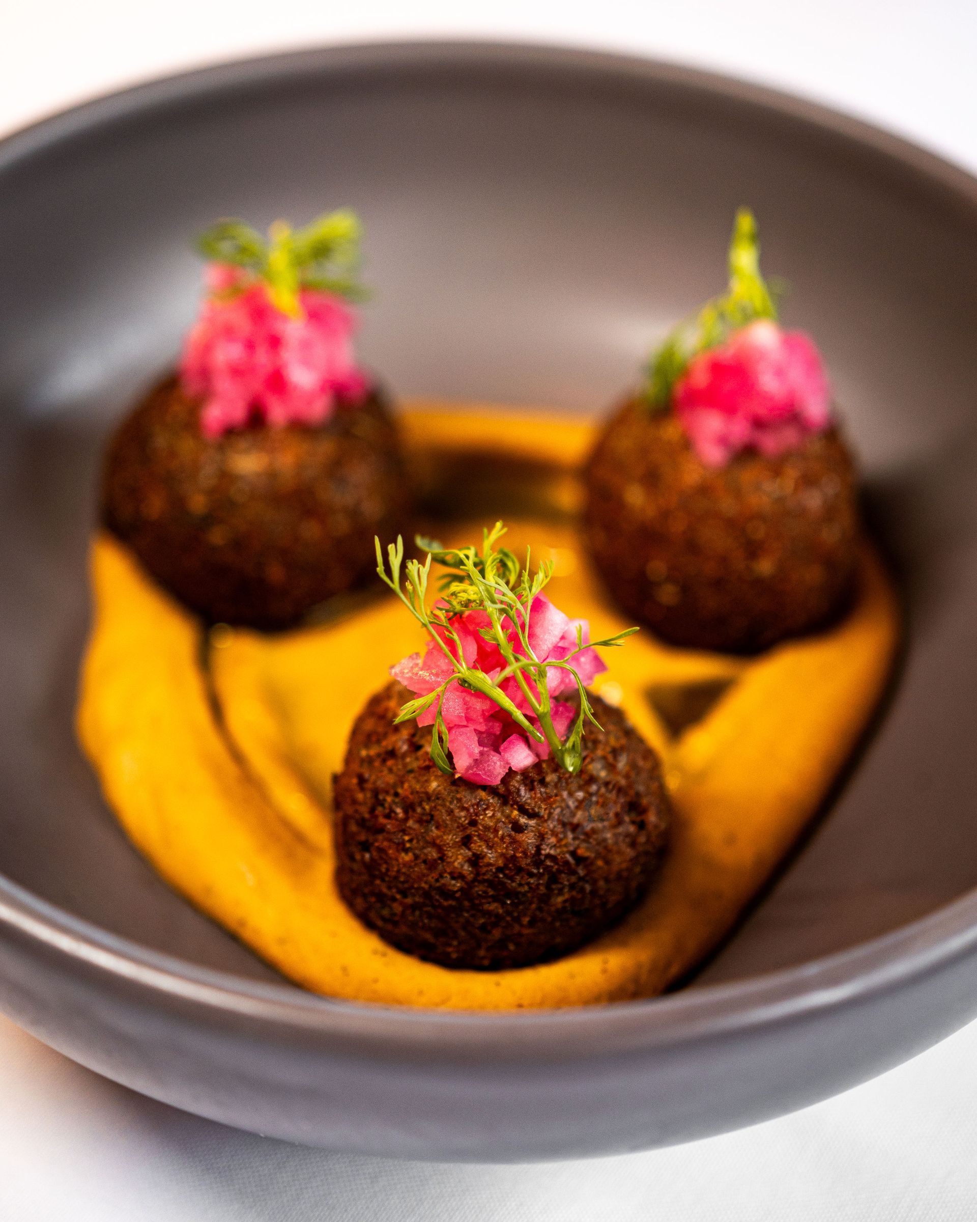 Three dark, round falafel balls topped with pink slaw on a bed of yellow sauce in a gray bowl.