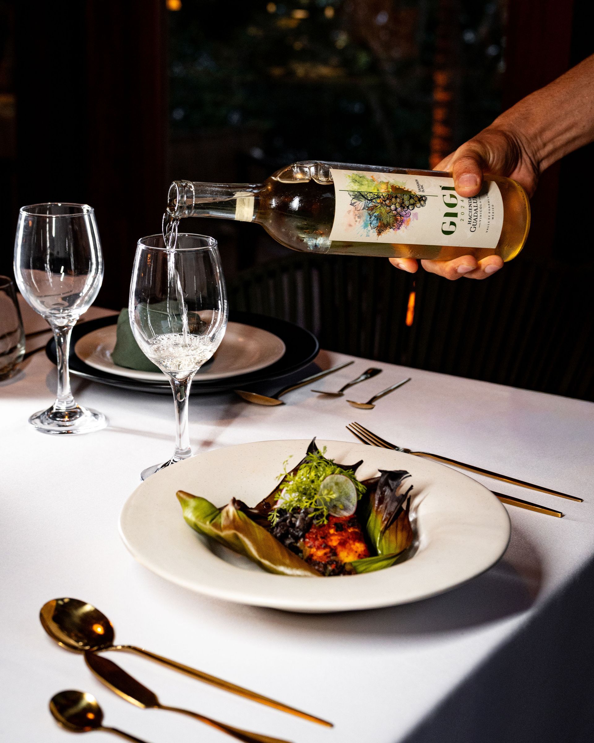 White wine being poured into a glass at a table set with food and gold utensils.