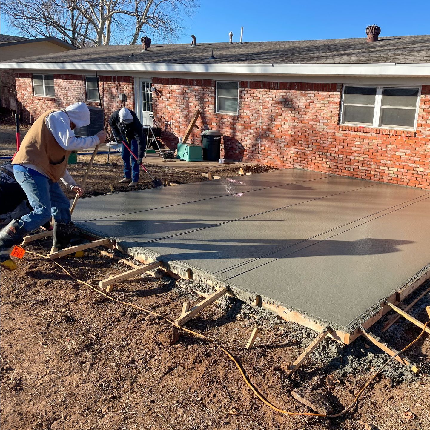 Construction workers pouring and leveling wet concrete for a patio near a red brick building.