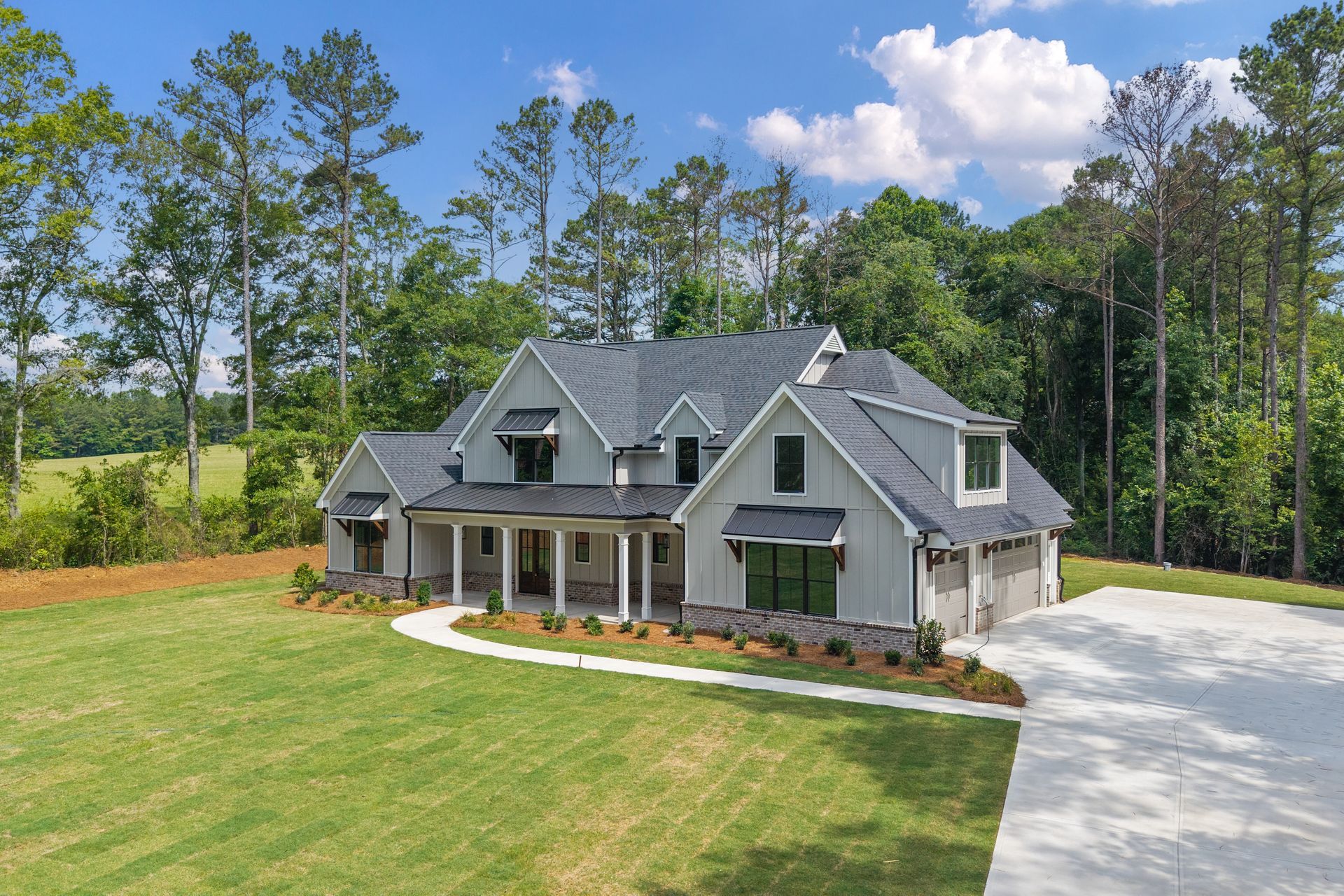 Gray farmhouse-style house with front porch, large lawn, and tree-lined driveway under a blue sky