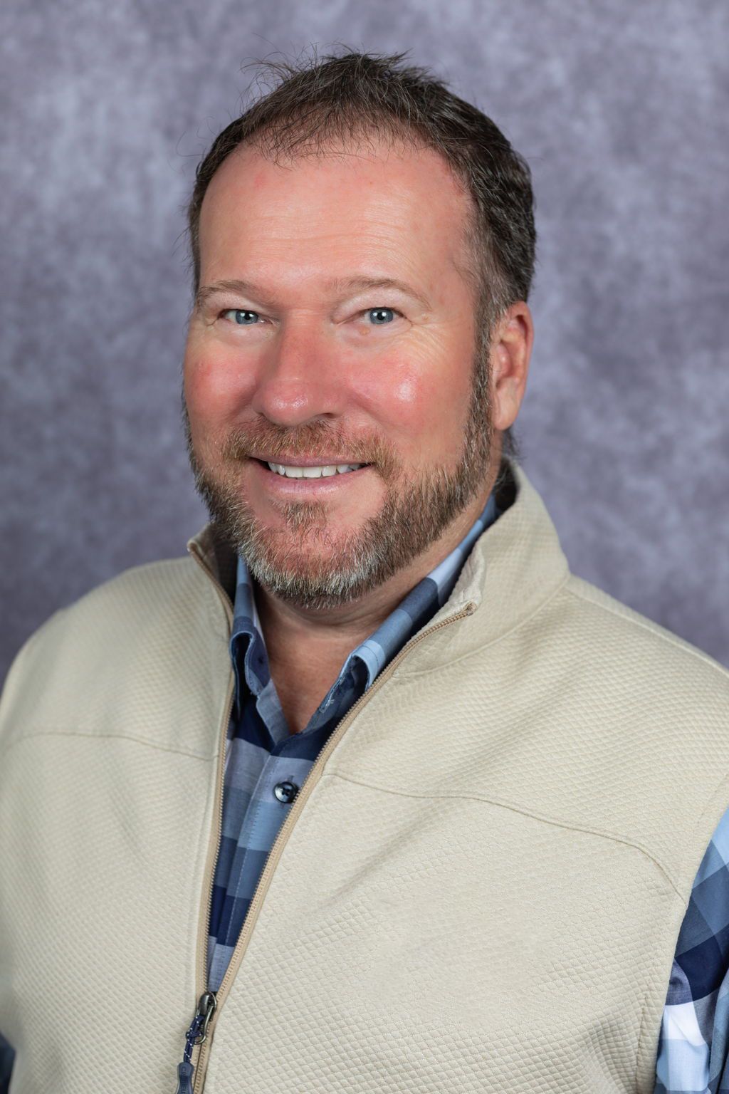 Man in plaid shirt and jeans smiles in front of a black fence, trees, and field.