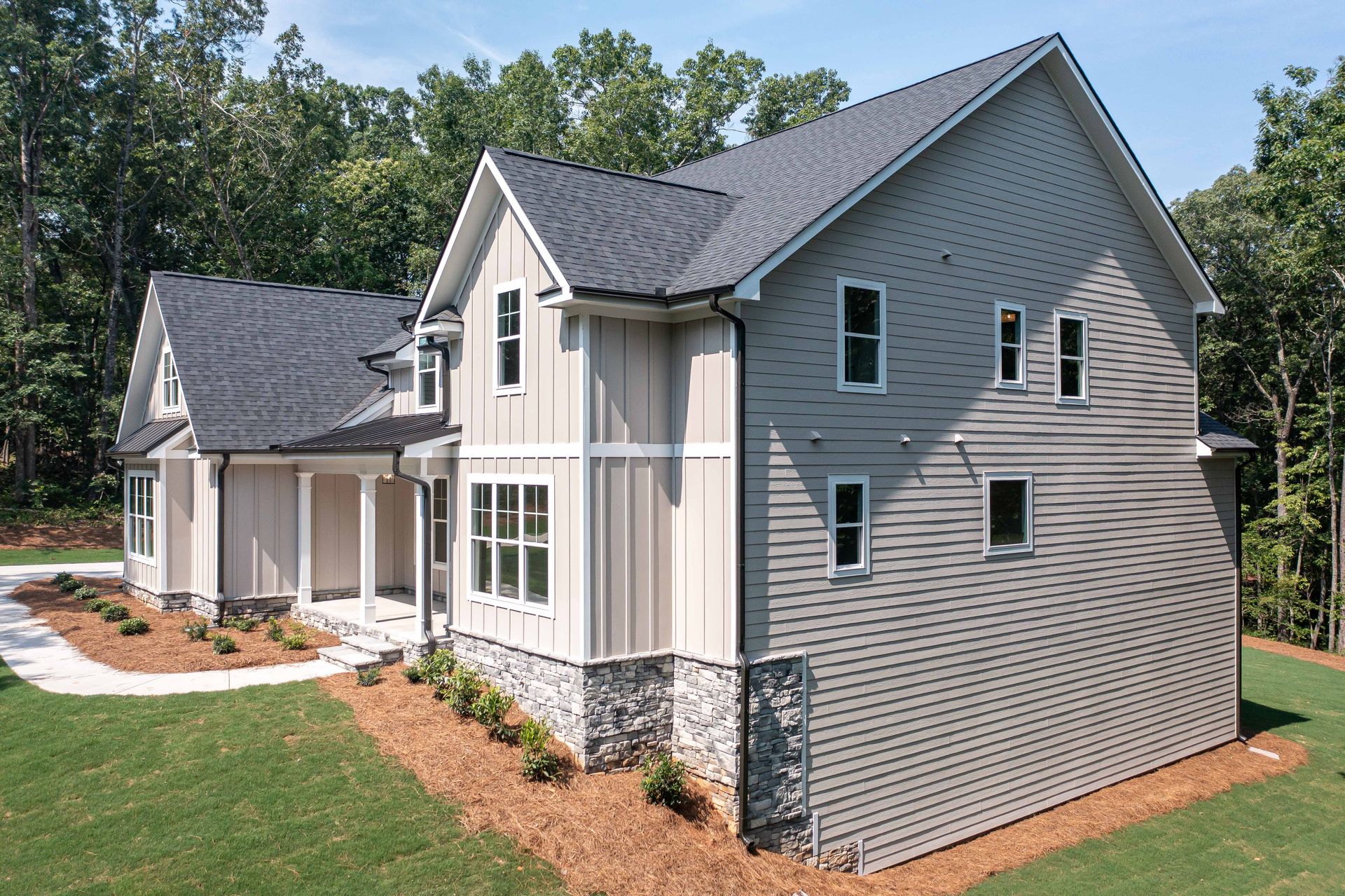 Modern two-story house with gray siding, black roof, and stone accents, surrounded by green grass and trees.