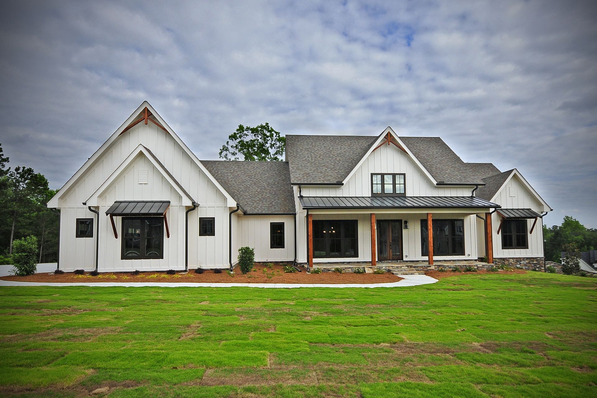 Two-story light gray house with black shutters, porch, and green lawn under a blue sky; another house in the background.