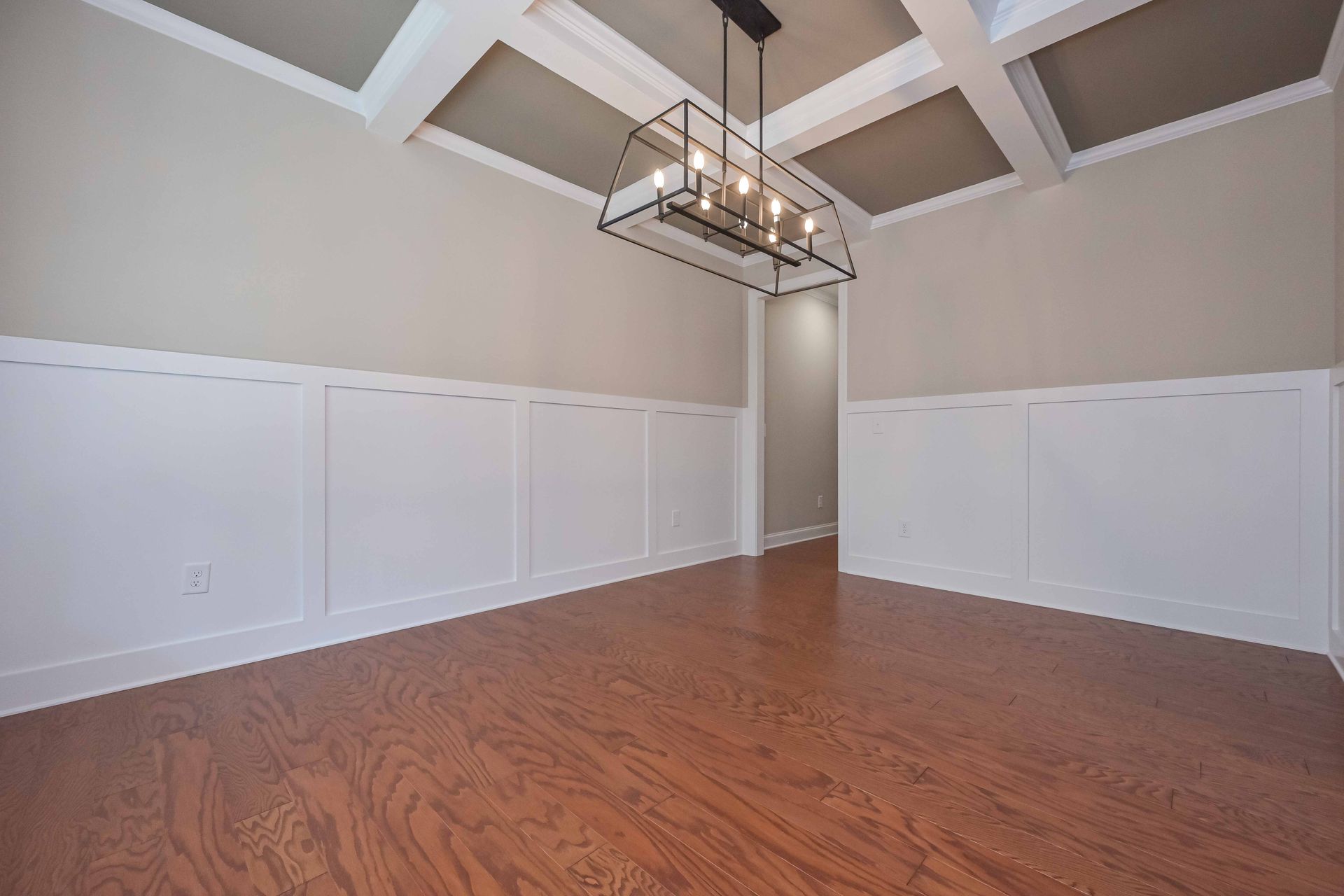Empty dining room with wood floor, white wainscoting, beige walls, and modern chandelier.