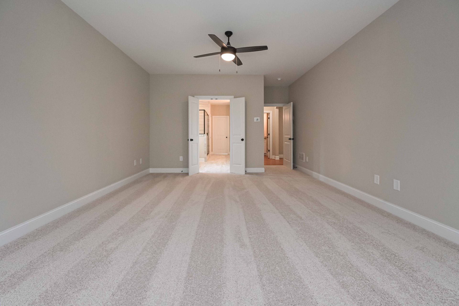Empty bedroom with neutral gray walls, carpet, ceiling fan, and doors leading to a bathroom.