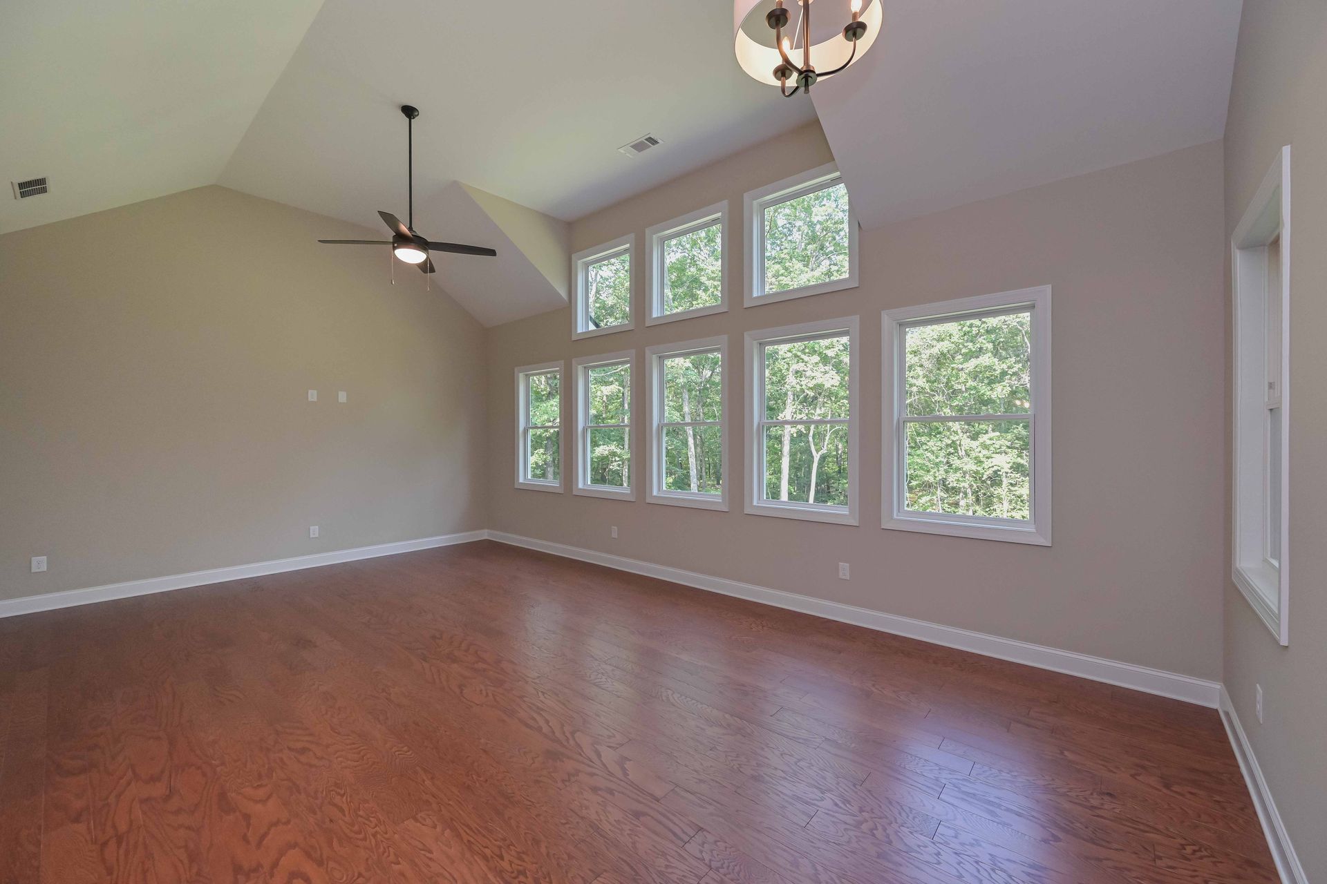 Empty room with hardwood floors, vaulted ceiling, and tall windows looking out to trees.