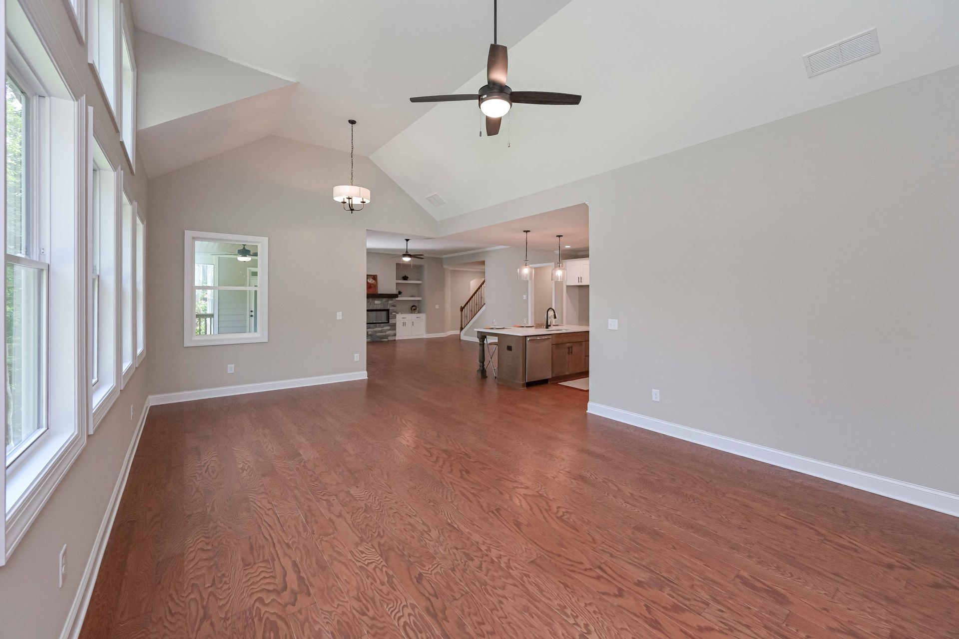 Empty living room with wood floor, high ceiling, windows, and view to the kitchen.