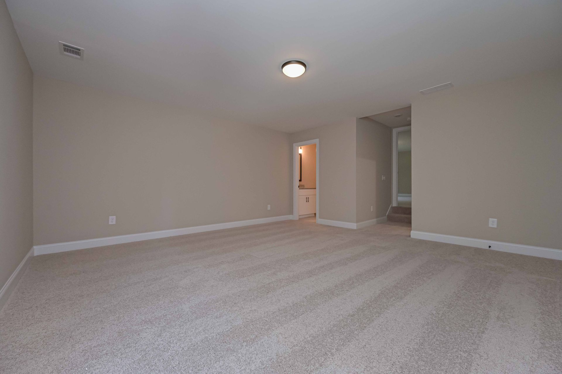 Empty room with beige carpet, walls, and trim. A doorway leads to a bathroom.