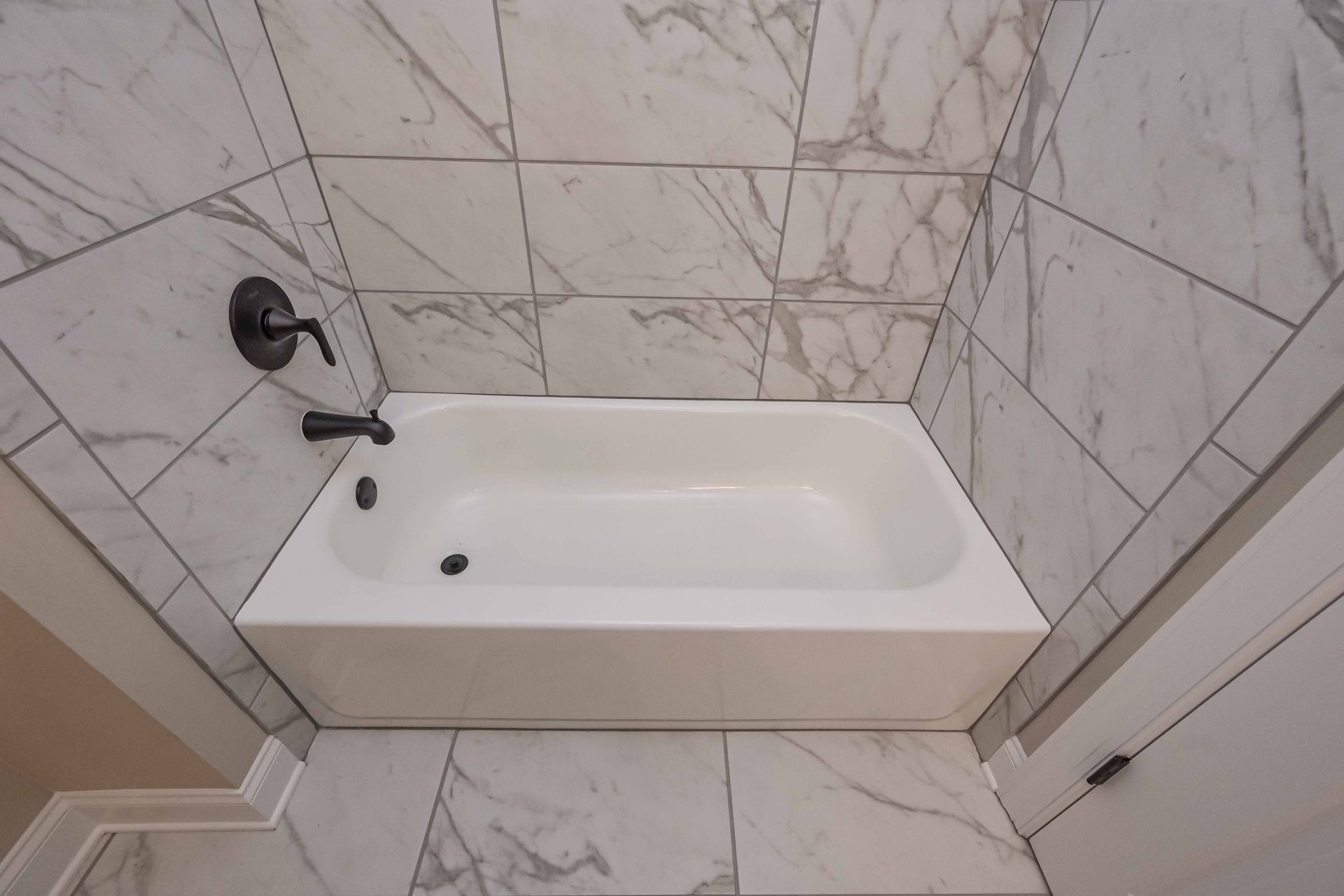 White bathtub with black fixtures, tiled surround with marble pattern.