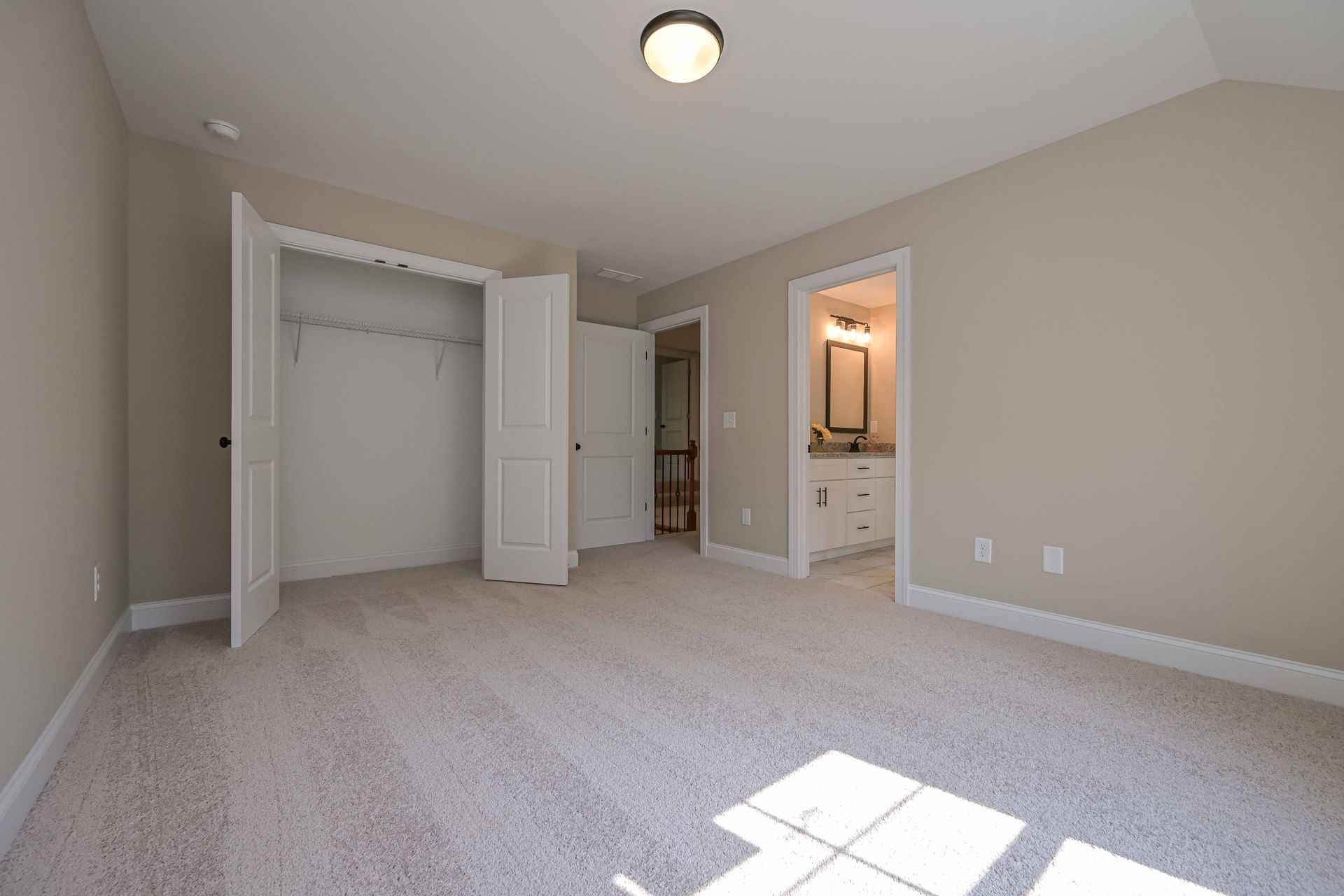 Empty bedroom with closet and bathroom door, tan walls, light carpet, overhead light.