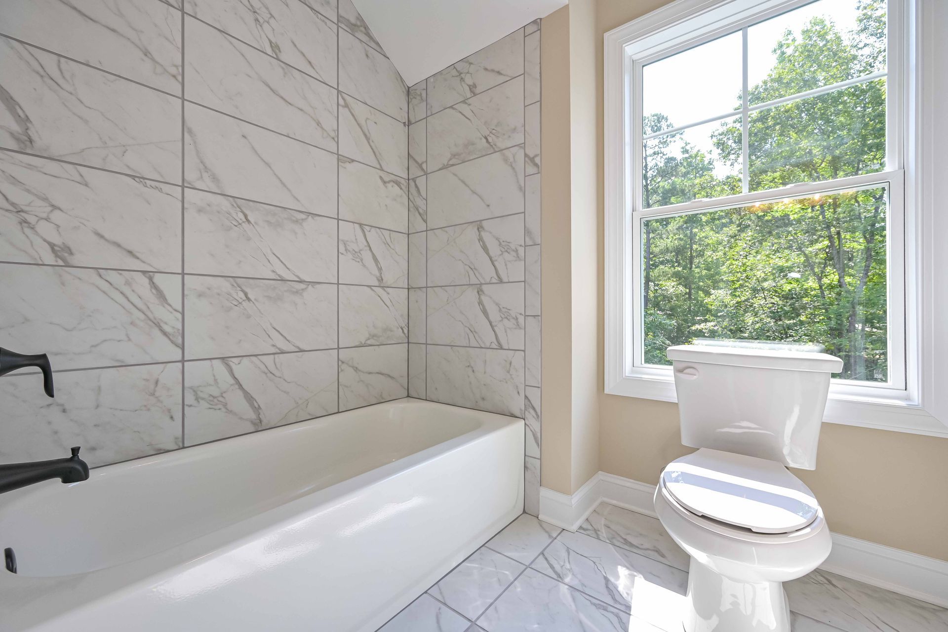 Bathroom with white tub, toilet, and marble tile, next to a window with trees outside.