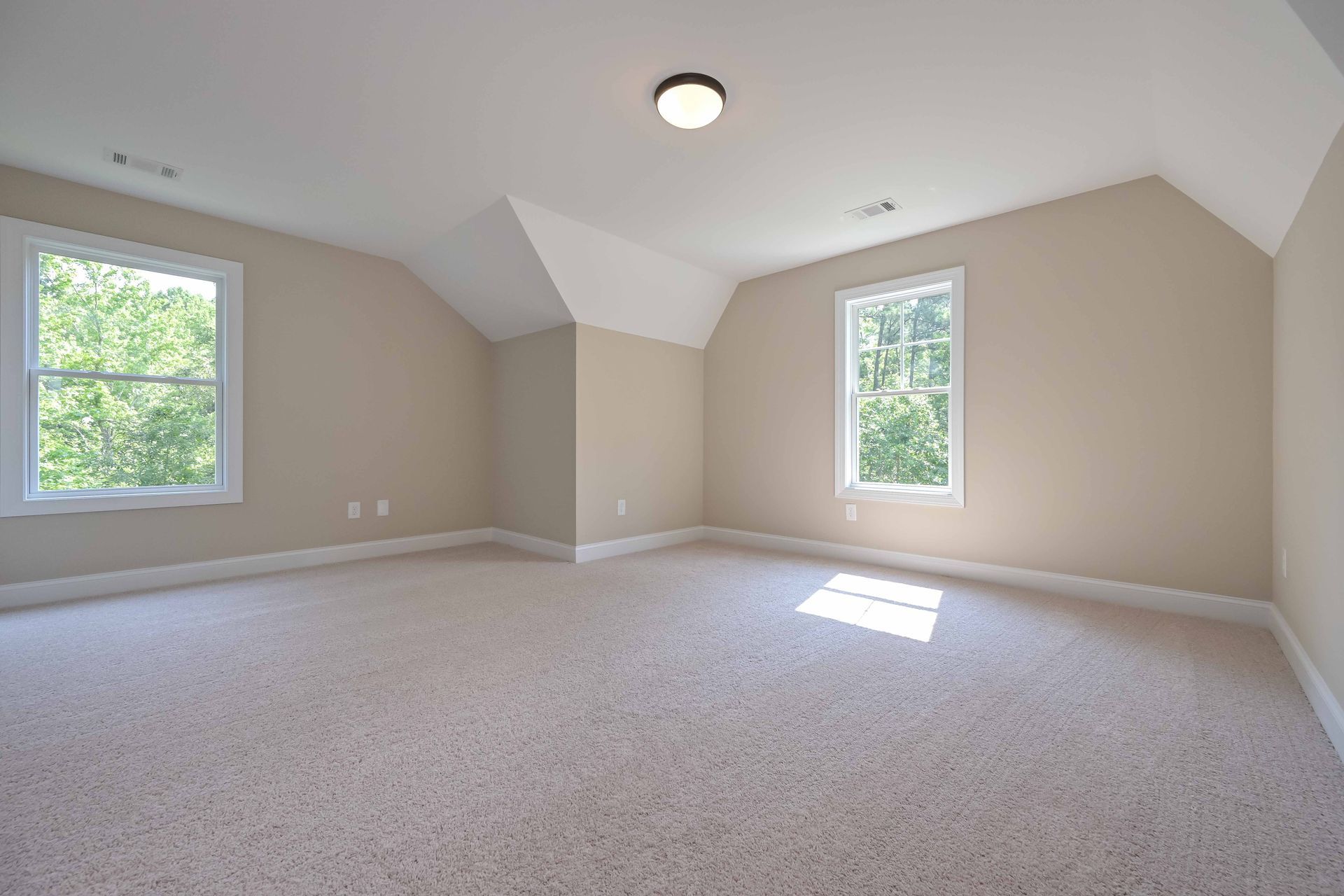 Empty beige carpeted room with two windows, a ceiling light, and white trim.