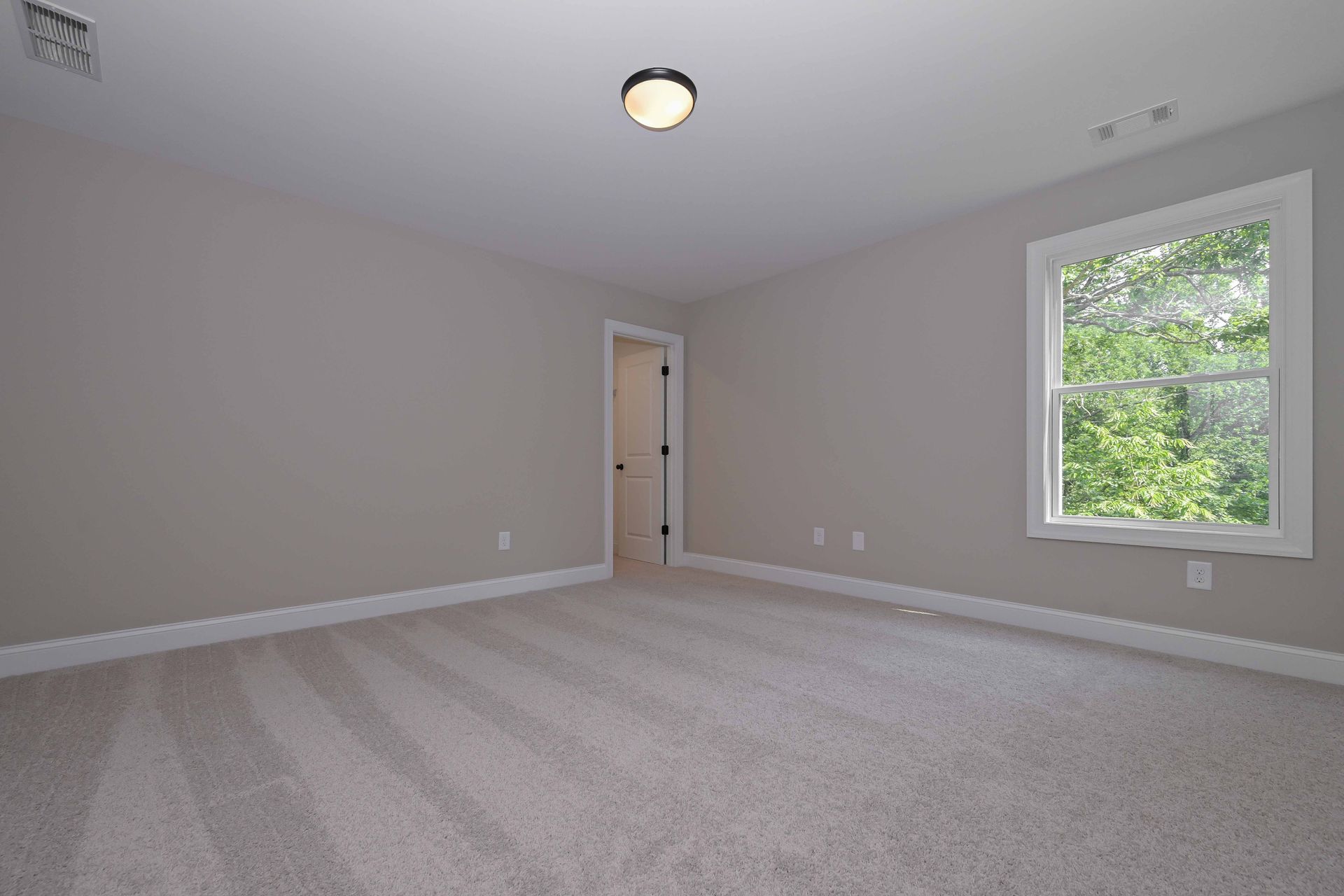 Empty bedroom with beige carpet, gray walls, and a window with a view of greenery.