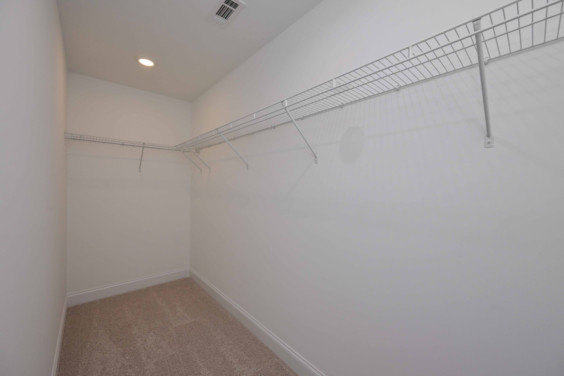 Empty walk-in closet with wire shelving and beige carpet. White walls, single ceiling light.