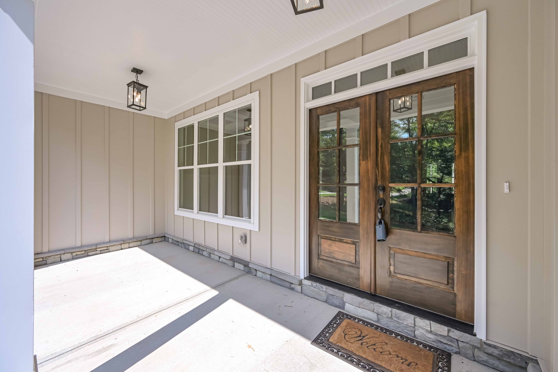 Covered porch with double wooden doors, window, tan siding, and concrete floor.