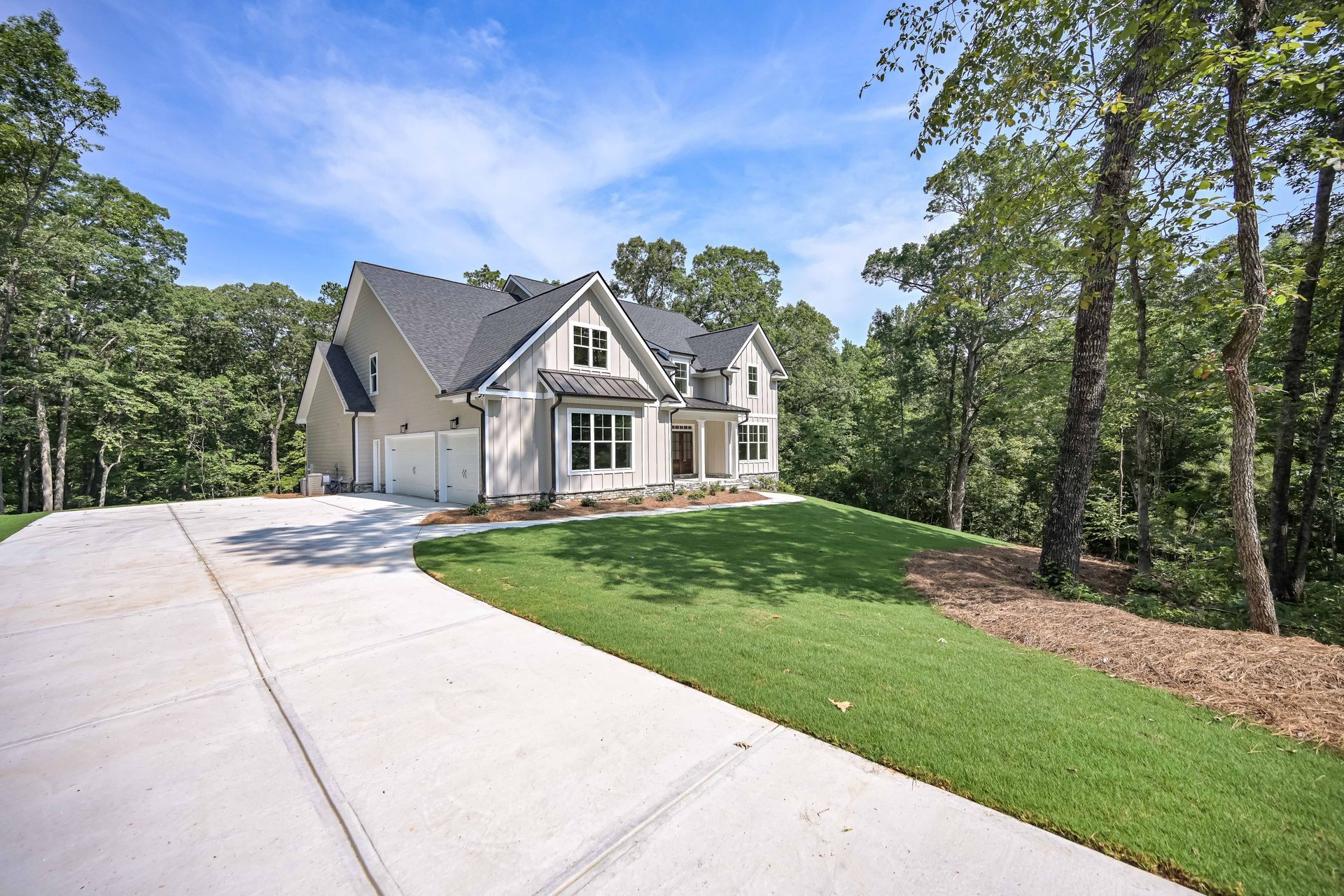 House with a concrete driveway, two-car garage, and green lawn, surrounded by trees under a blue sky.