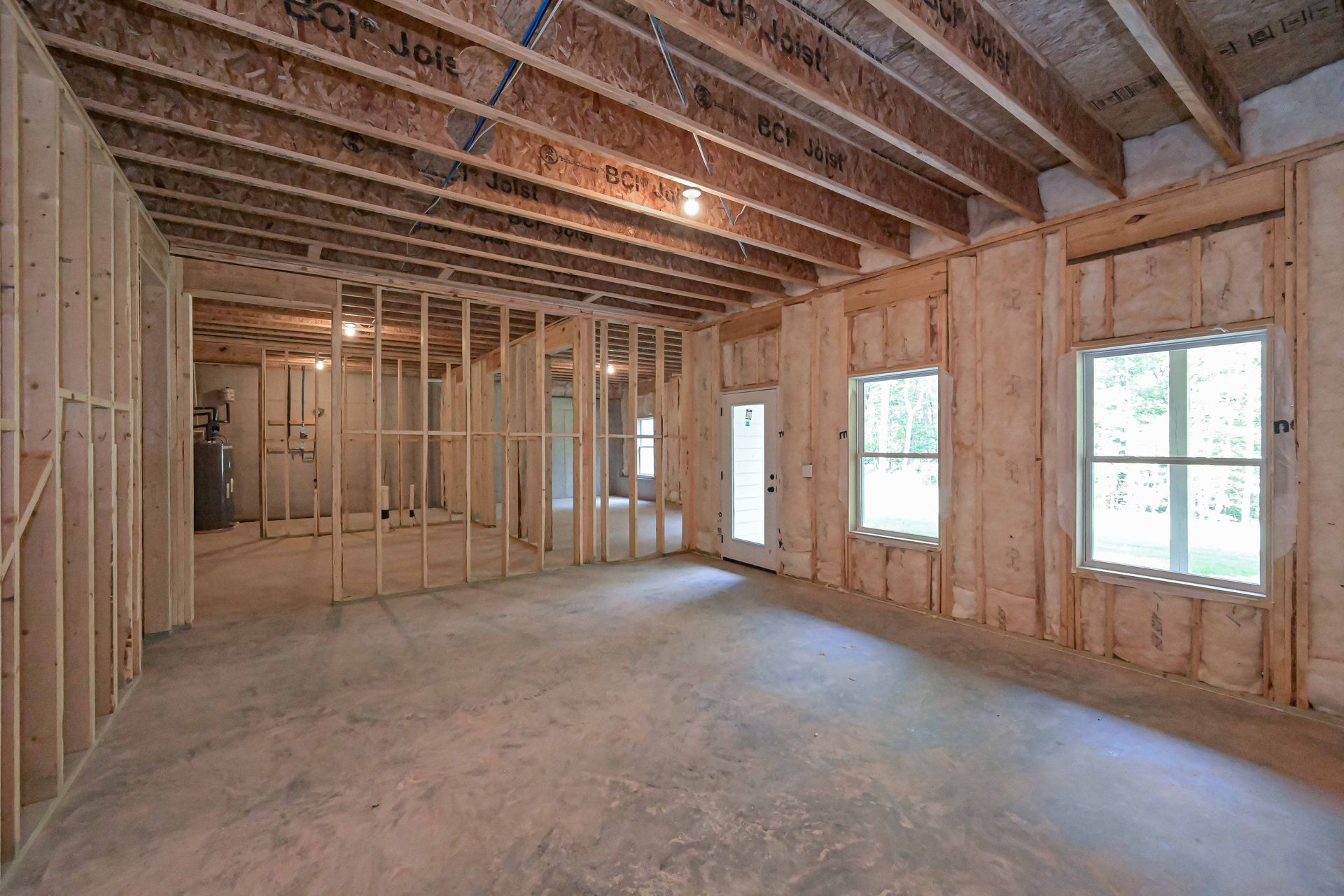 Interior of a home under construction. Exposed wooden framing, concrete floor, windows, and a doorway.